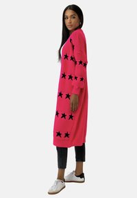 Long cardigan en tricot rose avec des motifs d'étoiles noires, des manches côtelées et des poches latérales. Associé à un jean noir et des baskets blanches.