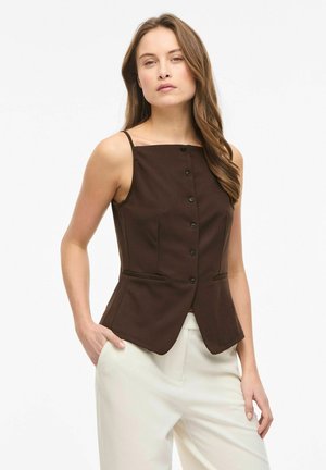 Femme aux longs cheveux bruns portant un haut sans manches marron foncé boutonné et un pantalon blanc, debout avec une main dans la poche.