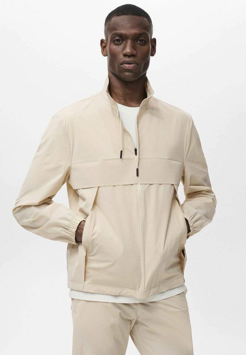 Beige zip-jacka i lättviktsmaterial med hög krage, elastiska muddar, två sidofickor och en färgblockeringsdesign.