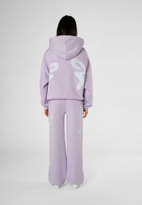 Personne portant un sweat à capuche violet clair et un pantalon large assorti avec des motifs abstraits blancs, debout de dos sur un fond blanc.