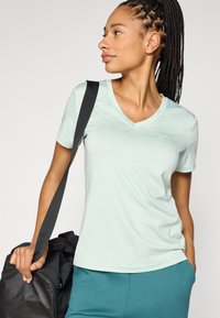 Ανοιχτό πράσινο-μέντα T-shirt με λαιμόκοψη σε V από μαλακό ύφασμα, κοντά μανίκια, συνδυασμένο με παντελόνια τιρκουάζ και μαύρη αθλητική τσάντα. Ομαλή υφή, άνετη εφαρμογή.