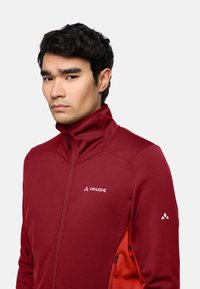 Rote Fleecejacke mit hohem Kragen, strukturiertem Material und seitlichen Reißverschlusstaschen. Mit einem weißen Logo auf der Brust und dem Ärmel.