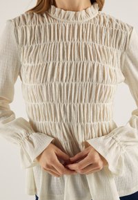 Bluza krem boje s teksturiranim, naboranim dizajnom i proširenim manšetama. Mekani, lagani materijal s detaljem ovratnika u naborima.
