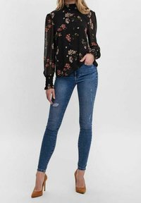 Blusa nera floreale con maniche lunghe, dotata di un collo alto e tessuto trasparente, abbinata a jeans skinny blu e tacchi alti color cammello.