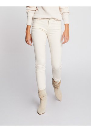 Persoon draagt offwhite slim-fit broek, beige losse enkellaarzen en een crèmekleurige gebreide trui tegen een effen lichte achtergrond.