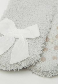 Zbliżenie na teksturowane jasnoszare podeszwy kapci, jedna z białą wstążką z materiału, a druga z małymi okrągłymi uchwytami na spodzie.