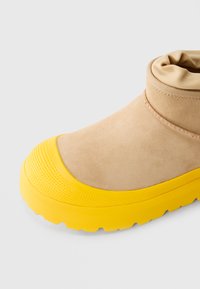 Cizme din suede bej cu vârf răsucit și o talpă groasă, texturată, de cauciuc galben strălucitor.