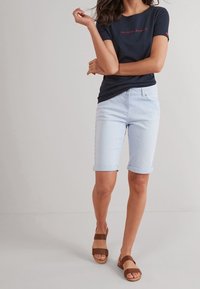 Granatowy t-shirt z czerwonym napisem; jasnoniebieskie szorty w paski z podwiniętymi mankietami; brązowe sandały na obcasie z dwoma regulowanymi paskami na gładkiej powierzchni.