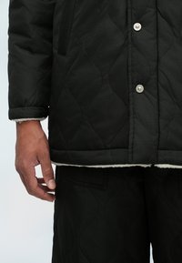 Giacca trapuntata nera con bottoni argentati, superficie testurizzata e colletto foderato in pile. Abbinata a pantaloni neri coordinati.