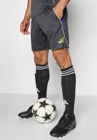 Shorts de sport gris avec des accents noirs et dorés, chaussettes noires avec des rayures blanches et chaussures de football noires, avec un ballon de football aux pieds.