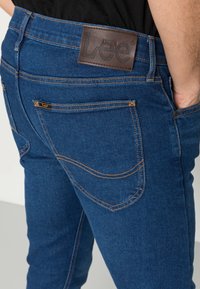 Os jeans de ganga azuis apresentam um rótulo de couro castanho com "Lee" em relevo na cintura, costura laranja e dois bolsos traseiros.