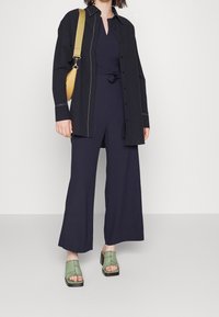 Žena v námořnickém overalu s uvázaným pasem, černou otevřenou košilí, zelenými sandály na podpatku a žlutou kabelkou na rameno na jednobarevném pozadí.