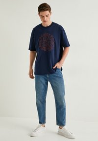 Marineblå bomulds t-shirt med korte ærmer, der har et cirkulært rødt grafisk tekstdesign. Kombineret med lyseblå denimjeans og hvide sneakers.