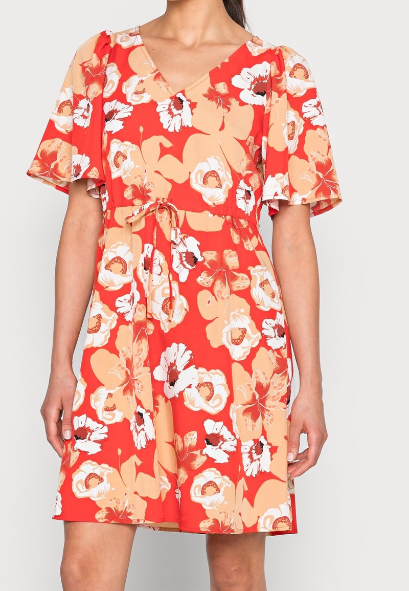 Femme portant une robe rouge à fleurs avec des fleurs pêche et blanches, des manches flottantes, un col en V et une ceinture nouée à la taille.