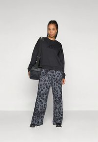 Μαύρη ζακέτα με υφασμάτινο λογότυπο, σε συνδυασμό με γκρι παντελόνι σε λεοπάρ σχέδιο με φαρδιά πόδια. Το μοντέλο κρατά μια μαύρη τσάντα ώμου. Μαύρα αθλητικά παπούτσια στα πόδια.