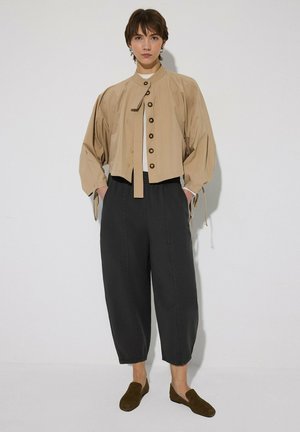 Personne debout contre un mur blanc portant une veste beige boutonnée, un pantalon noir ample et court, des chaussures marron sans lacets, et les mains dans les poches.