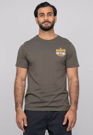 Homme aux cheveux foncés et à la barbe, portant un t-shirt vert olive avec un petit motif jaune d'Indianapolis sur la poitrine gauche, debout devant un fond uni.