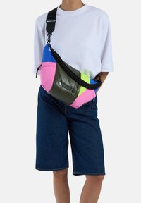 Sac banane avec un mélange de matériaux bleus, roses, verts et olive. Surface texturée avec des fermetures éclair et une bandoulière tressée. Design compact et fonctionnel.