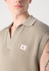 Bež pletena brezrokavna polo majica z V-izrezom, z rebrasto teksturo in logotipom na levi strani.