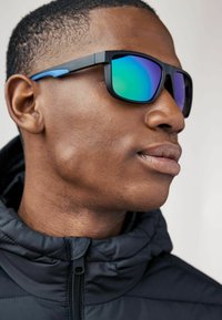 Ochelari de soare sport cu ramă neagră, accente albastre și lentile verzi reflectorizante. Purtați cu o geacă neagră căptușită.