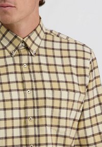 Korte mouwen, lichtgele geruite shirt met bruine ruitpatronen, knoopsluiting en een puntige kraag met decoratieve knopen.