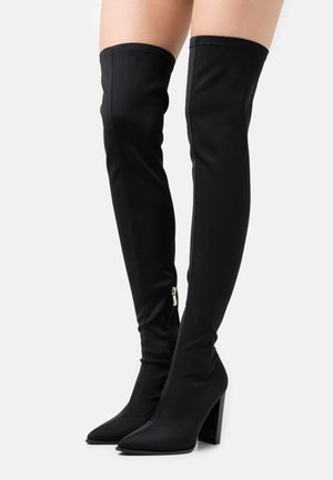 Bottes noires montantes en tissu extensible. Présentent un bout pointu et un talon carré, avec un zip sur le côté pour un enfilage facile.