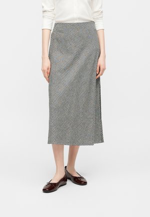 Kobieta ubrana w czarno-białą, kraciastą midi spódnicę, białą koszulę z długim rękawem oraz ciemnobrązowe baleriny z lakierowanej skóry.