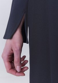 Dunkelgraues Kleid aus strukturiertem Stoff, mit einem seitlichen Schlitz und langen Ärmeln. Eine Hand ruht sanft auf dem Stoff in der Nähe des Ärmelausschnitts.