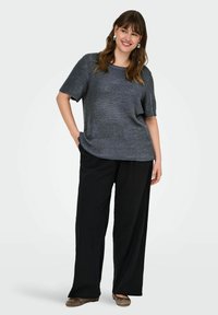 Szary dzianinowy top z krótkim rękawem o luźnym kroju, zestawiony z luźnymi czarnymi spodniami, tworząc casualowy zestaw. Brązowe baleriny dopełniają stylizację.