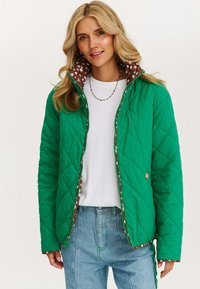 Kobieta w zielonej pikowanej kurtce z brązowym wzorzystym podbiciem, białej koszuli, niebieskich dżinsach i kolorowej naszyjniku z koralików, uśmiechająca się do kamery.