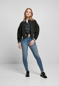 Musta bomber-takki valkoisilla raidoilla hihojen resoreissa ja kauluksessa, kerrospukeutuneena vihreän ruudullisen paidan päälle, yhdistettynä vaaleansinisiin skinny-farkkuihin ja mustiin kenkiin.