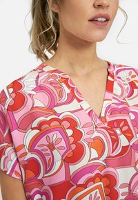 Bluzka z krótkim rękawem z dekoltem w szpic w geometryczny różowo-czerwony wzór z kwiatowymi akcentami i gładką teksturą materiału.