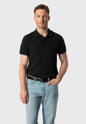 Hombre con cabello corto y rizado, vestido con una camiseta polo negra y vaqueros azul claro, de pie con una mano en el bolsillo frente a un fondo liso.
