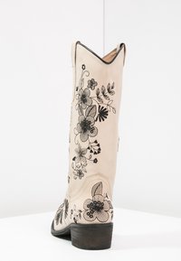 Stivale da cowboy in pelle beige con ricami floreali neri sul gambale, punta affusolata e tacco nero impilato, con un bordo superiore leggermente ondulato.