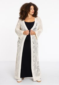 Langer, offener Strickcardigan in Creme, mit einem Rautenmuster, lockeren Ärmeln und einem fließenden Design, getragen über einem schwarzen Kleid.