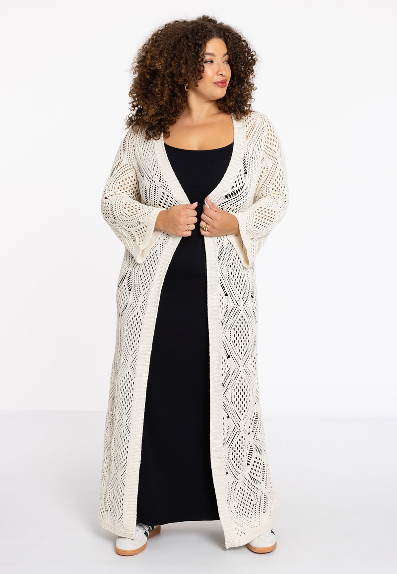 Lange, opengehaakte cardigan in crème, met een ruitpatroon, losse mouwen en een vloeiende uitstraling, gedragen over een zwarte jurk.