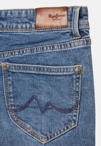 Vaqueros de mezclilla azul con un parche de cuero marrón en la cintura y un patrón de olas cosido en el bolsillo trasero.