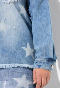 Chaqueta de mezclilla azul claro con patrones de estrellas blancas, detalles con tachuelas, dobladillo deshilachado y cierre de botón en el puño.