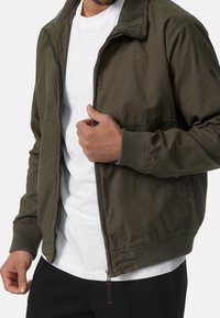 Veste bomber vert olive avec fermeture éclair, poignets et col côtelés. Présente un logo discret et deux poches latérales, fabriquée en tissu léger.