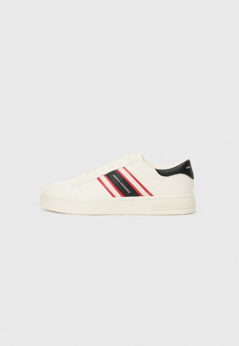Cream-colored sneakers feature three red and black stripes, a black heel accent, and a textured rubber sole. Smooth fabric and lace-up design.