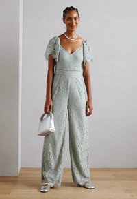 Spetsöverdel i ljus mintgrön med korta genomskinliga puffärmar, v-ringning, vida ben och blommönster. Accessoarer med en silverhandväska.
