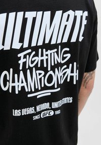 Camiseta negra con texto blanco que dice "Ultimate Fighting Championship, Las Vegas, Nevada, Estados Unidos, desde UFC 1993" y un brazo tatuado.