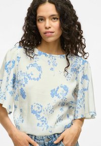 Blusa di crema chiara con motivo floreale blu, maniche corte, scollo rotondo, tessuto morbido e vestibilità comoda, abbinata a pantaloni shorts in denim.