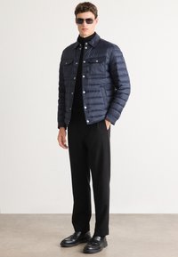 Moški v temnomo modri zloženi jakni, črnem turtlenecku, črnih hlačah, črnih loaferjih in sončnih očalih, stoji pred enobarvno belo ozadjem.