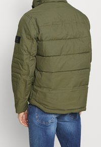 Uomo che indossa un giubbotto imbottito a forma di piumino verde oliva e jeans blu, visto di spalle su uno sfondo chiaro e uniforme.