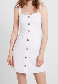 Weißes Leinenkleid mit tailliertem Oberteil, quadratischem Ausschnitt und breiten Trägern. Verfügt über Frontknöpfe in Goldfarbe und einen ausgestellten Rock.