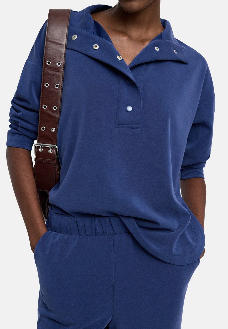 Granatowy sweter z kołnierzykiem, zapinany na zatrzaski, z długimi rękawami. Tkanina wydaje się miękka, o luźnym kroju. Połączony z dopasowanymi niebieskimi spodniami.