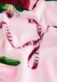 Coperta in pile con uno sfondo rosa e motivi floreali. Include un nastro di raso con la scritta "BAKER" stampata in lettere scure. Texture morbida.