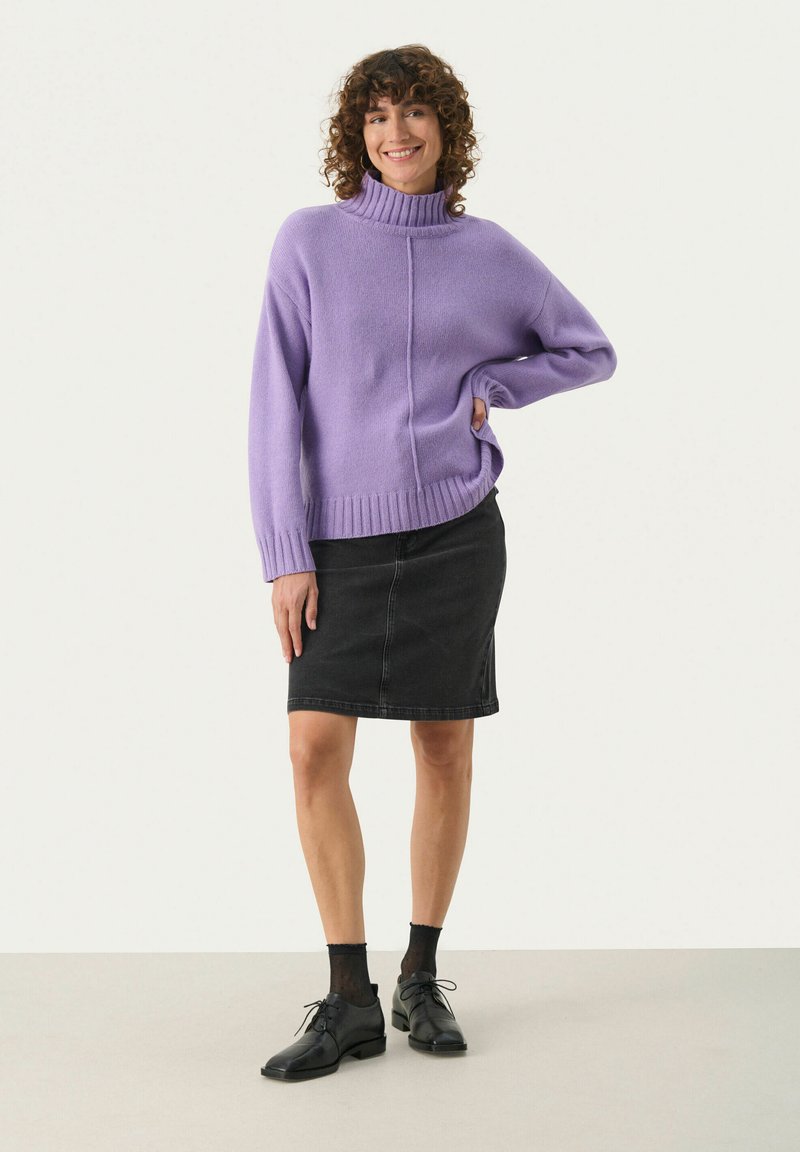 Liliowy sweter z golfem, z prążkowanymi mankietami i dołem, zestawiony z czarną spódnicą dżinsową. Modelka ma na sobie czarne buty i wzorzyste skarpetki.