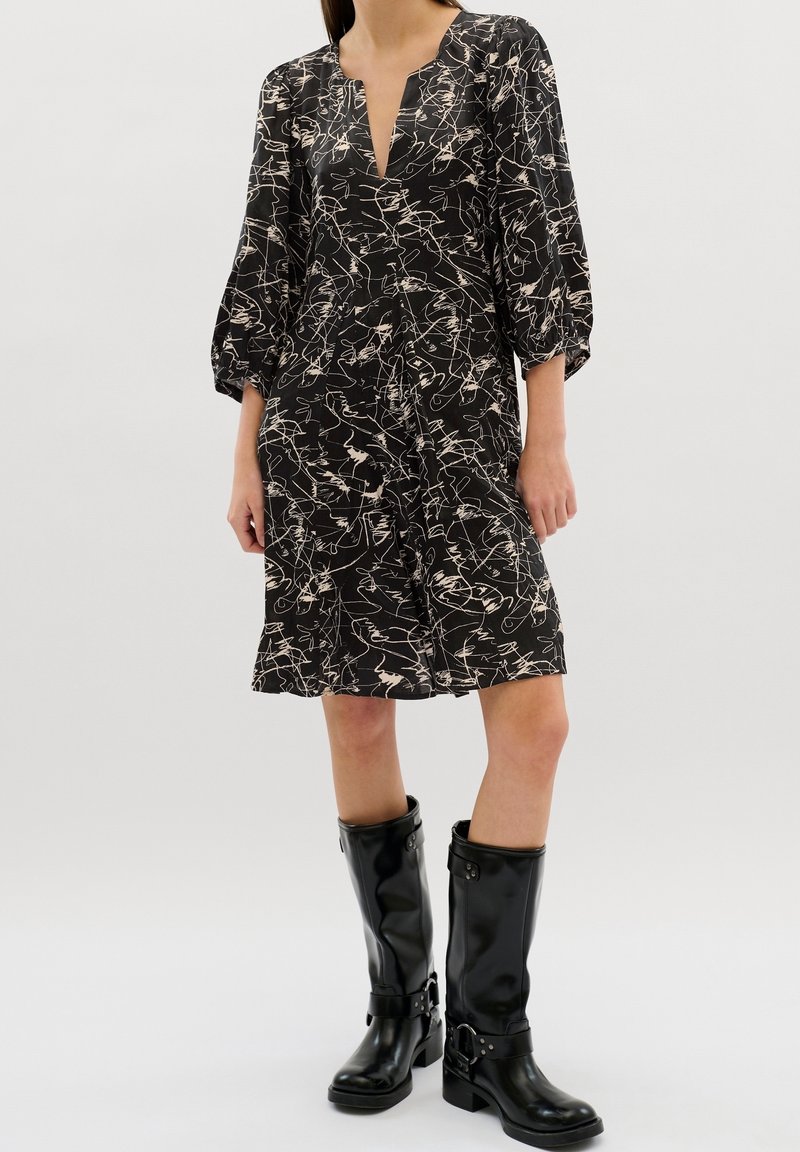 Femme portant une robe noire longueur genou avec des motifs abstraits en lignes blanches et des bottes en cuir noires mi-mollet sur un fond clair.
